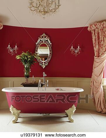 Stock Image - Traditional red and white bathroom with rolltop bath and venetion mirror and chandelier.. Fotosearch