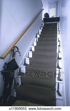 Stock Image - Umbrellas and collection of old iron shoe lasts on steep white staircase with grey carpet and wooden handrail. Fotosearch