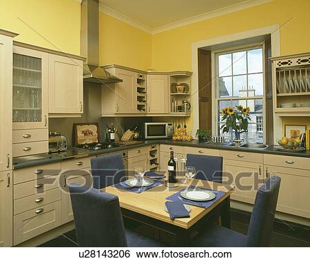 Stock Photograph - Upholstered blue chairs and wooden table in pastel yellow kitchen with cream fitted cupboards. Fotosearch