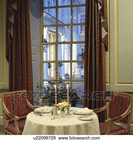 Upholstered chairs and circular table with place settings on white cloth in front of tall window with brown curtains View Large Photo Image Stock Photo - Upholstered chairs and circular table with place settings on white cloth in front of tall window with brown curtains. Fotosearch