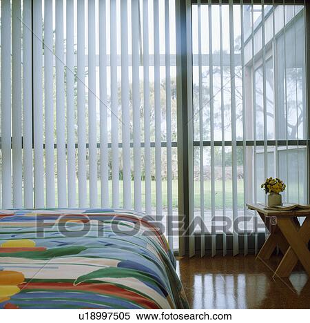 Stock Photography - Vertical white slatted blinds at large windows in modern bedroom with multicoloured duvet. Fotosearch