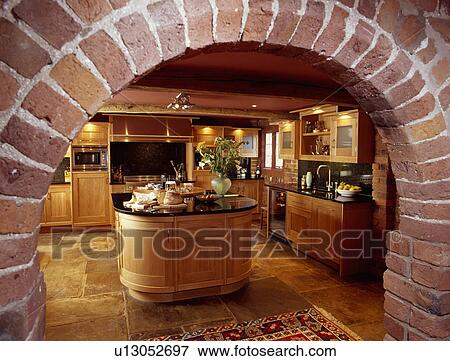 Stock Photo - View through brick arch of traditional wooden kitchen with oval island unit and stone flooring. Fotosearch