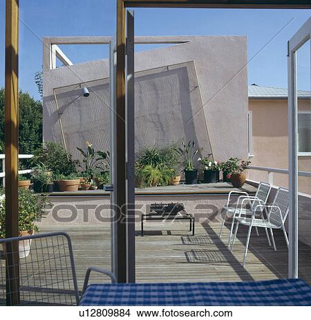 View through glass doors of dining room to decked city roof terrace with barbeque and white chairs and plants in pots View Large Photo Image Picture - View through glass doors of dining room to decked city roof terrace with barbeque and white chairs and plants in pots. Fotosearch
