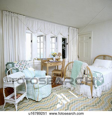 Stock Image - White curtains and striped turquoise armchair in coastal bedroom with ziz-zag striped carpet and single wooden bed. Fotosearch