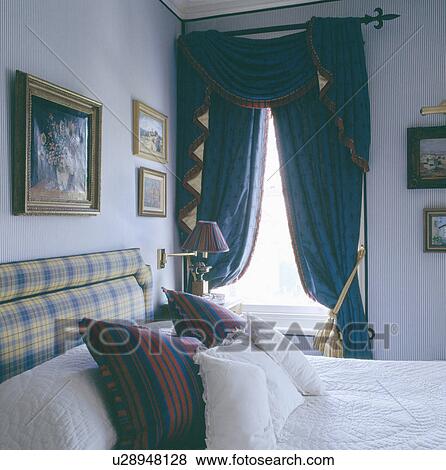 White cushions on bed with upholstered headboard in grey bedroom with blue swagged and tailed curtains View Large Photo Image Stock Photo - White cushions on bed with upholstered headboard in grey bedroom with blue swagged and tailed curtains. Fotosearch