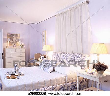 White Drapes Above Blue White Upholstered Bed With White Bolster Pillow In Mauve Bedroom With Lighted Lamps Stock Photography