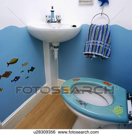 Stock Photograph - White pedestal basin in corner of bathroom with painted trompe-l'oiel seascape and blue novelty fish-pattern toilet-seat. Fotosearch