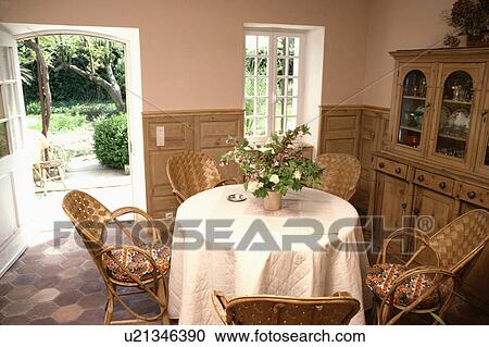 Stock Image - White quilted cloth on circular table with woven rattan chairs in country dining room with open door to garden. Fotosearch