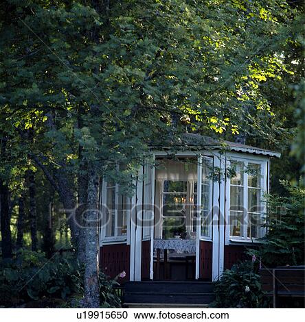 Stock Image - White wooden summerhouse below trees in large country garden in summer. Fotosearch