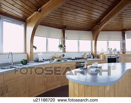 Stock Image - Wooden ceiling in circular pale wood modern kitchen with island unit and white blinds on row of windows and. Fotosearch