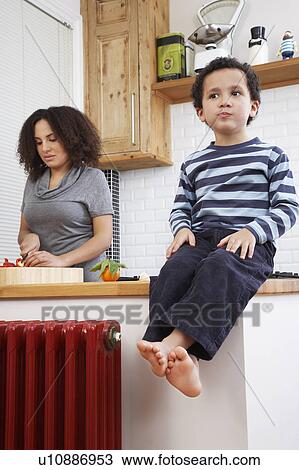 Stock Image - Boy (5-6) sitting on kitchen counter, mother in backbround. Fotosearch