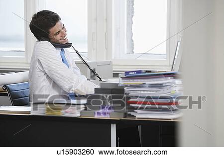 Stock Image - Business man using telephone at desk in office, smiling. Fotosearch