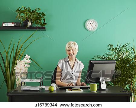 Picture - Business woman behind desk in office full of potted plants, portrait. Fotosearch