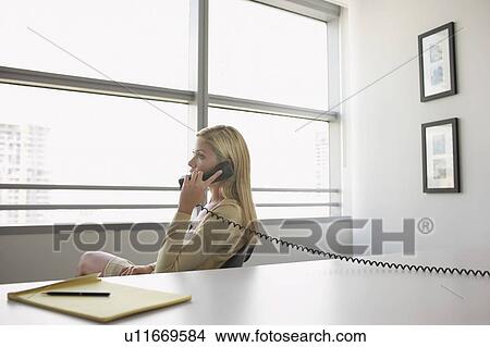 Picture - Business woman using phone sitting at office desk, side view. Fotosearch