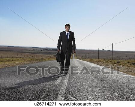 Stock Image - Businessman walking down road in isolated landscape. Fotosearch