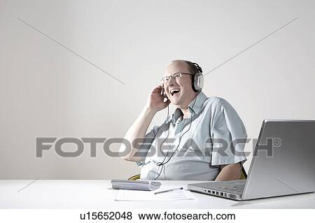 Stock Photo - Businessman with earphones laughing at desk. Fotosearch