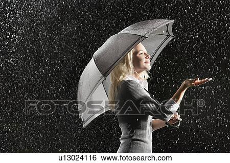 Stock Photograph - Businesswoman sticking hand out from under umbrella during rain, side view. Fotosearch