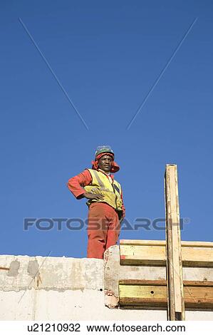 Stock Image - Construction worker stands with hands on hips on incomplete building. Fotosearch