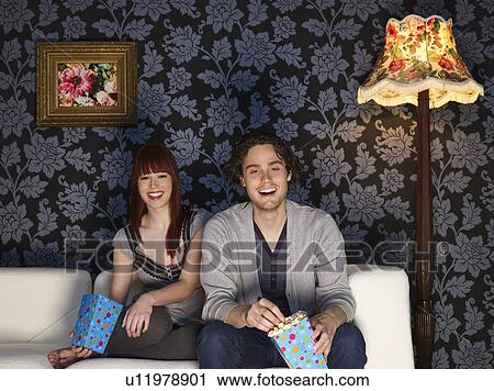 Couple sitting on sofa holding popcorn bags, laughing View Large Photo Image Stock Image - Couple sitting on sofa holding popcorn bags, laughing. Fotosearch