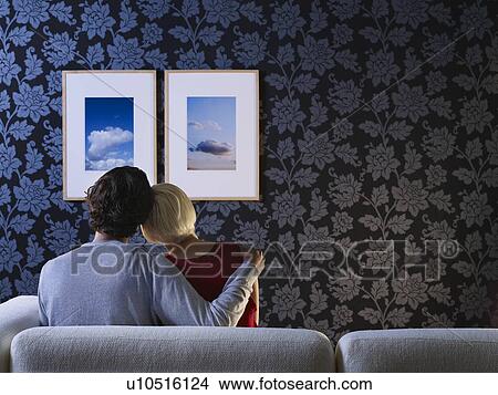 Picture - Couple sitting on sofa looking at pictures on wall, back view. Fotosearch