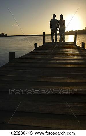 Stock Image - Couple standing on dock by lake, holding hands, back view.. Fotosearch