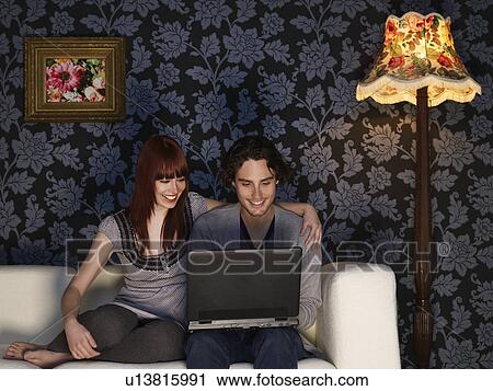 Stock Image - Couple using laptop sitting on sofa. Fotosearch