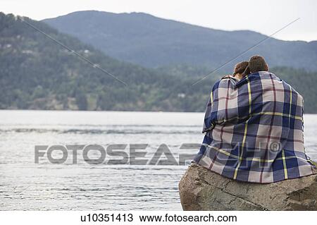 Stock Image - Couple wrapping in blanket, sitting on rock by ocean. Fotosearch