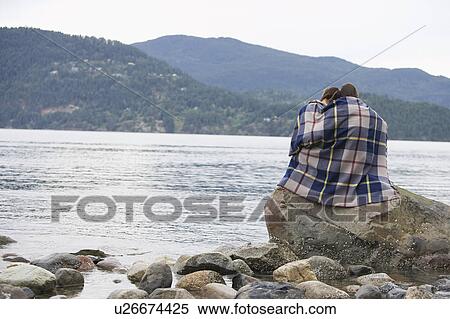 Stock Photography - Couple wrapping in blanket, sitting on rock by ocean. Fotosearch