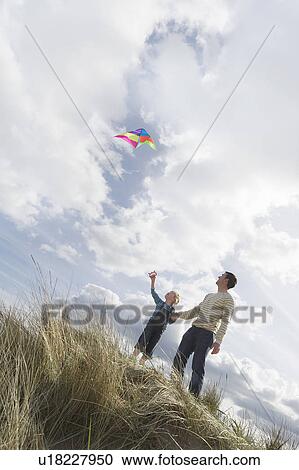 Stock Image - Father and daughter (5-6) flying kite on dunes. Fotosearch