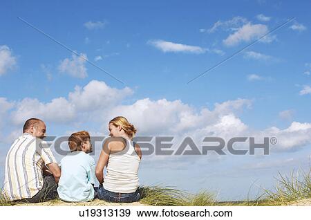 Stock Photo - Father and mother sitting with son on sand dune, back view. Fotosearch