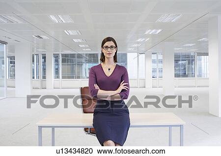Stock Image - Female office worker sitting on edge of table in empty office space. Fotosearch
