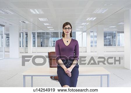 Stock Photo - Female office worker sitting on edge of table in empty office space. Fotosearch