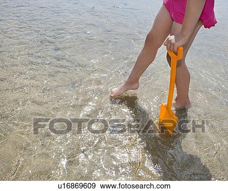 Stock Photo - Girl (10-12) with toy spade in shallow water, low section. Fotosearch