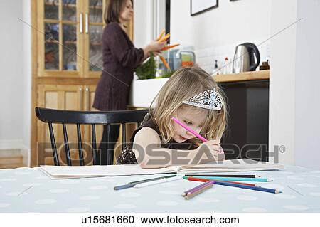 Stock Image - Girl (3-4) drawing in kitchen, mother preparing food in background. Fotosearch