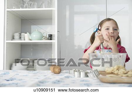 Stock Image - Girl (5-6) preparing food in bowl, using wire whisk and licking finger. Fotosearch