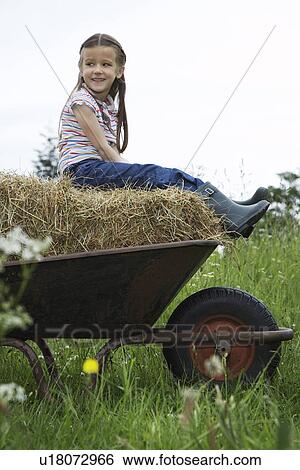 Stock Photograph - Girl (5-6) sitting on hay in wheelbarrow in field, portrait. Fotosearch