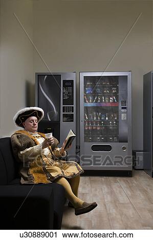Stock Image - King Henry VIII reading on sofa near vending machine. Fotosearch