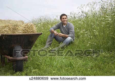 Stock Photo - Man sitting beside wheelbarrow with hay in field, portrait. Fotosearch