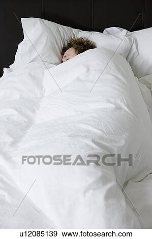 Stock Photo - Man sleeping in bed covered under duvet, elevated view. Fotosearch