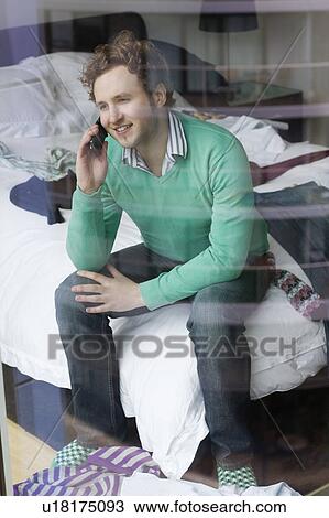 Stock Image - Man using mobile phone sitting on bed, view through window. Fotosearch