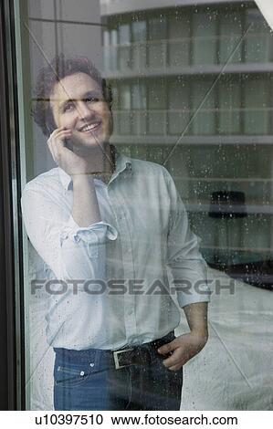 Stock Image - Man using mobile phone standing by window. Fotosearch