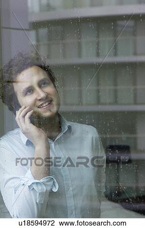 Stock Image - Man using mobile phone standing by window. Fotosearch