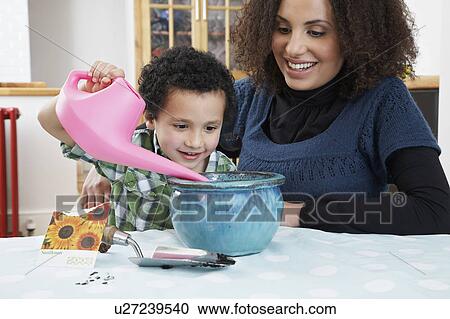 Stock Image - Mother assisting son (5-6) pouring water into flower pot. Fotosearch