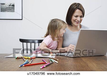 Stock Photograph - Mother sitting at table with daughter (3-4), using laptop, smiling. Fotosearch