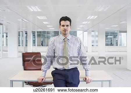 Stock Image - Office worker perching on edge of table in empty office space. Fotosearch