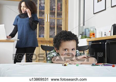 Stock Image - Portrait of boy (5-6) sitting at table, mother in background using laptop and phone. Fotosearch