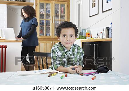 Stock Image - Portrait of boy (5-6) sitting at table, mother using laptop in background. Fotosearch