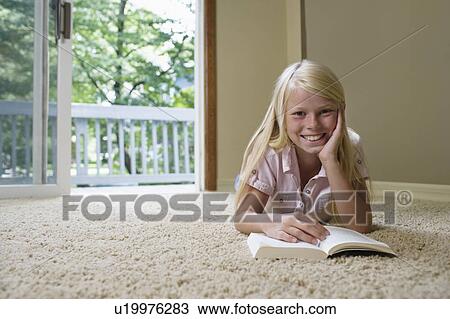Stock Image - Portrait of girl (7-9) reading book on carpet, smiling. Fotosearch