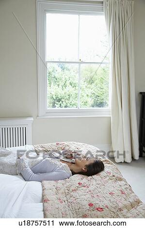 Stock Image - Portrait of young woman lying on bed talking on phone. Fotosearch