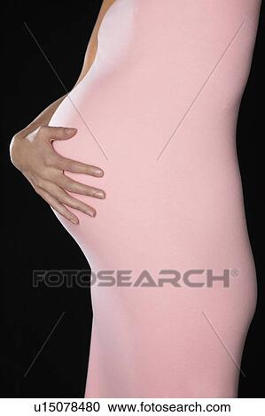 Stock Image - Pregnant woman in body suit holding abdomen, mid section, side view, studio shot. Fotosearch